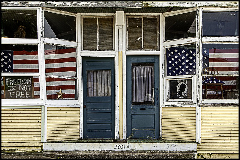 vintage store front with yellow-painted siding and big windows displaying American flags, a POW MIA sign and a chalkboard with freedom is not free written on it