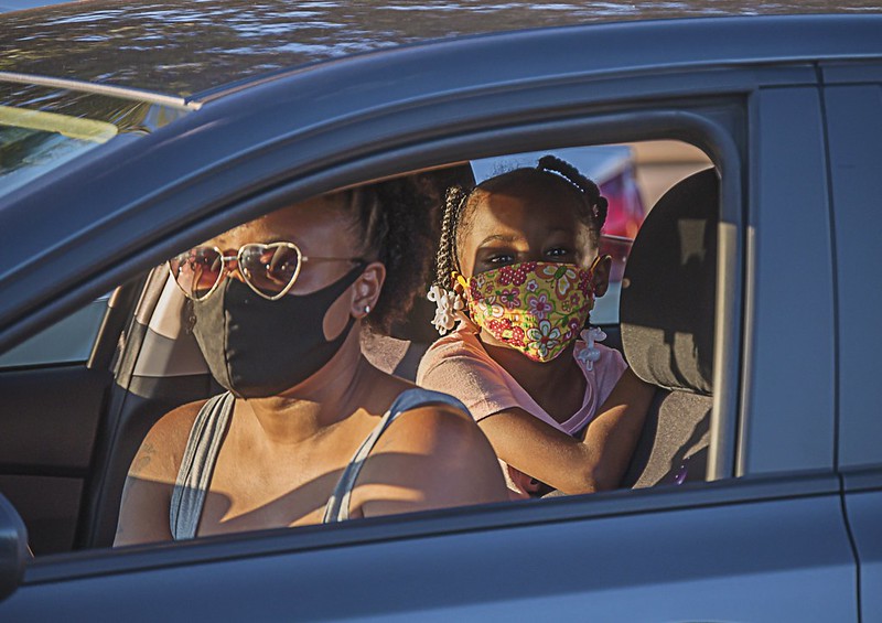 a young African-American woman wearing heart-shaped sunglasses and a medical mask driving a car with her daughter in the backseat, who is also wearing a mask