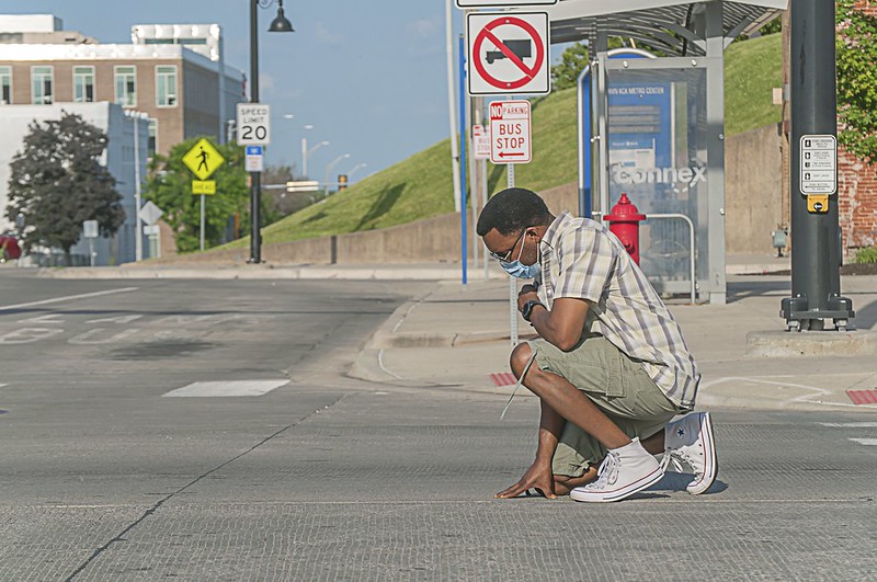 an African-American man wearing a medical mask kneeling in prayer in the middle of an urban stree intersection